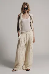 Pantalón de tiro medio confeccionado en hilatura con lino, con cintura con trabillas, bolsillos laterales ocultos y pierna ancha. Presenta un diseño con estampado jacquard de motivos florales en tonos beige y crudo, y cierre frontal con cremallera, botón y ganchos metálicos.