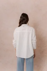 Camisa de denim azul con bordados florales en tonos amarillos y celestes sobre los hombros y el pecho. Presenta cuello clásico, cierre frontal con botones y mangas largas con puños.