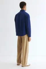 Sobrecamisa azul de lona gruesa de algodón, con cuello, tapeta con botones a presión y canesú en la espalda. Tiene mangas largas con botón a presión en los puños y dos bolsillos superiores de parche. El bajo es levemente redondeado y el corte es oversized.