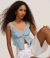 Top cropped de jeans celeste con escote corazón, breteles anchos y abertura frontal central abotonada. Los bordes del escote y la abertura están decorados con un ribete de encaje blanco tipo picot.