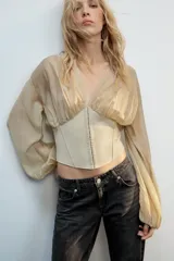 Top beige de manga larga con escote en pico, mangas abullonadas y puños elásticos. Presenta un diseño combinado con tejido fluido en la parte superior y tejido estructurado tipo corsé en la cintura, con cierre frontal de ganchos metálicos.