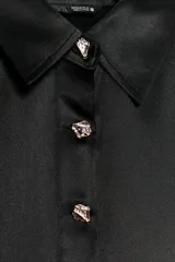 Camisa de satén color blanco, de corte clásico con cuello solapa, manga larga con puños y cierre frontal con botones dorados.