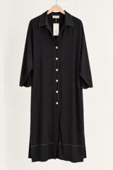 Vestido midi negro con diseño camisero, cuello camisero, cierre frontal con botones blancos y costuras a contratono. Cuenta con mangas tres cuartos y aberturas laterales en el dobladillo.