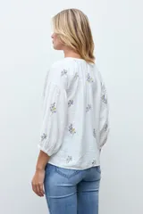 Blusa blanca de corte holgado con escote en V y mangas tres cuartos abullonadas. Presenta un delicado bordado de pequeñas flores en tonos azules y amarillos dispersas por toda la prenda.