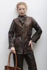 Blazer de cuero negro con silueta recta y estructura sastrera, cierre frontal, cuello alto y múltiples bolsillos con tapa y herrajes. Cuenta con un recorte triangular en el pecho.