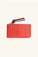 Monedero rectangular de cuero vacuno con textura granulada en color marrón topo. Cuenta con cierre superior metálico y un tirador decorativo de cuero en color naranja con un nudo. Presenta un pequeño logo grabado en el centro.