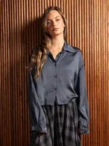 Camisa de raso color gris oscuro, con cuello clásico, cierre frontal de botones y mangas largas con puños abotonados. Presenta un corte corto y holgado.