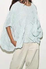 Top de cuello redondo color celeste con textura fruncida, mangas tipo capa y bajo asimétrico.