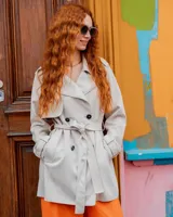 Campera trench de gabardina en color beige, con solapa cruzada, doble botonadura frontal, hombreras y cinturón ajustable para anudar en la cintura. Presenta bolsillos delanteros y puños ajustables con tiras.