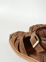 Sandalias estilo cangrejera color marrón, confeccionadas en cuero con diseño trenzado en la parte delantera, tira ajustable con hebilla metálica en el tobillo y suela plana.