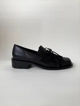 Mocasín de cuero marrón con textura granulada, diseño de horma cuadrada, detalle de flecos y lazo decorativo en el empeine, montado sobre una suela y taco bajo de color negro.