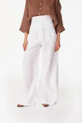 Pantalón de lino blanco de corte ancho y fluido, con cintura alta que incorpora una sobrepretina ancha desmontable con pliegues horizontales.