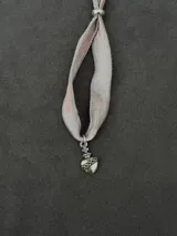Colgante con cinta de tela grisácea y rosada, adornado con un dije de corazón sagrado plateado y cruz.