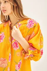 Camisa de viscosa con estampado floral en tonos amarillos y rosas. Presenta escote redondo con cierre frontal de botones, mangas al codo con terminación elástica y detalle de volado.