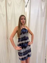 Vestido corto con escote halter y cuello alto, con tiras ajustables en el cuello. Presenta un diseño estampado con efecto tie-dye en tonos azul, negro y blanco, con espalda abierta y elástico en la cintura.