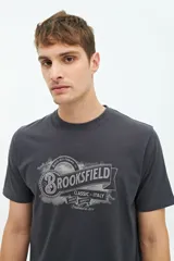 Remera de manga corta color beige con estampado gráfico frontal de la marca Brooksfield en tonos grisáceos, con diseño de estilo vintage y tipografía clásica.
