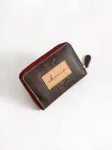 Billetera pequeña de cuero color chocolate con cierre perimetral rojo y detalle de costuras decorativas en forma de espiga en el frente.
