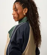 Campera rompeviento de diseño tricolor con bloques de color en verde, beige y blanco. Presenta cuello alto, cierre frontal con cremallera, mangas raglán y puños ajustados. El ruedo incluye un cordón regulable para un ajuste personalizado.