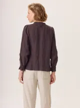 Camisa de lino rústico color marrón oscuro, con cuello mao, botones frontales y detalles de volados en el frente. Mangas largas con puños fruncidos.