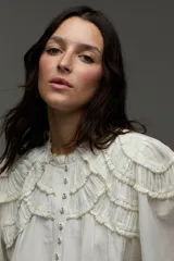 Blusa blanca con cuello mao, detalles de mini volados y bordados en el pecho y las mangas, con cierre frontal de botones y mangas largas abullonadas.