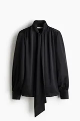 Blusa de gasa negra con cuello alto y lazada frontal, mangas largas abullonadas con puños abotonados y detalle de frunces en los hombros.
