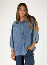 Camisa de denim azul con cuello camisero, cierre frontal de botones y mangas 3/4 abullonadas con puños elásticos.