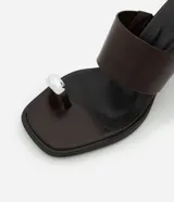 Sandalia tipo mule color marrón oscuro, con punta cuadrada, tacón alto y una tira ancha en el empeine. Presenta un detalle de dedera metálica plateada en la parte frontal.