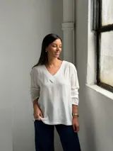 Blusa blanca de corte holgado con escote en V, mangas largas y costuras visibles en el cuello y el ruedo.