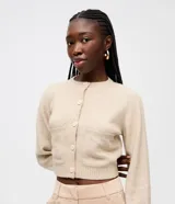 Cardigan corto de punto color beige con textura atoallada, cuello redondo, manga larga y cierre frontal de botones. Cuenta con dos bolsillos delanteros y detalles acanalados en el bajo y los puños.