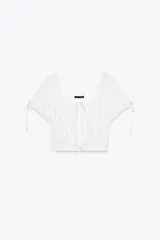 Top corto blanco de gasa con escote cuadrado, mangas cortas y bordados de flores en el frente. Presenta lazada a contraste en escote y mangas, y cierre frontal con botones.