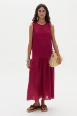 Vestido midi de lino color beige, sin mangas, con cuello redondo y detalles de ondas en las sisas. Presenta un corte con volante fruncido en la parte inferior.