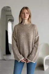 Sweater de tejido de punto en color beige con textura suave y detalles de costuras visibles en color blanco en los hombros, puños y ruedo. Presenta un diseño de corte holgado, cuello redondo y mangas largas.