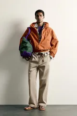 Cazadora de corte relajado para hombre, confeccionada en algodón con acabado efecto encerado en color marrón. Cuenta con cuello subido, capucha ajustable con cordones, mangas largas con puños elásticos y cierre frontal con cremallera. Incluye bolsillos frontales de vivo y forro interior a cuadros.