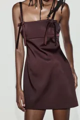 Vestido mini confeccionado en tejido satinado color bordó, de escote recto con tirantes finos y detalle de lazos aplicados en el delantero.