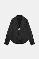 Camisa de satén negro con diseño jacquard de flores tono sobre tono. Presenta escote en pico con solapas, cierre frontal con botones y un broche metálico decorativo en el centro. Tiene mangas largas con puños abotonados.