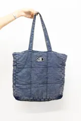 Bolso tipo tote confeccionado en tela denim acolchada con diseño de costuras cuadradas. Presenta doble asa superior y un parche ovalado con el logo 'dl' en el frente.