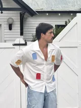Camisa blanca de manga corta con parches de tela de colores (azul, rojo, amarillo) distribuidos irregularmente.