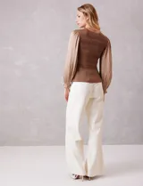 Blusa de punto con torso texturizado en color marrón con hilos de lurex dorados. Presenta un corte peplum con volado en la cintura y mangas largas de satén fluido con volumen, terminadas en puños elásticos fruncidos.
