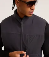 Chaleco deportivo gris oscuro con cuello alto y cierre frontal con cremallera. Presenta recortes en tejido liso en la parte superior del pecho y en los bolsillos laterales.