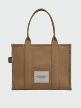 Cartera tipo tote bag Marc Jacobs modelo The Large Tote, color marrón con el texto "THE TOTE BAG MARC JACOBS" impreso en blanco en el frente. Confeccionada en lona resistente, cuenta con asas superiores dobles para llevar al hombro o en la mano.