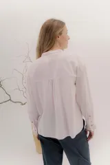 Camisa de algodón blanca con cuello a la base, cierre frontal con botones y mangas largas con puños delicados. Presenta volados verticales y detalles de cintas de terciopelo en contraste a lo largo del frente.