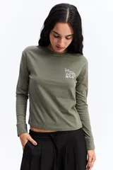 Remera de manga larga color verde oliva, con cuello redondo y estampa gráfica pequeña en el pecho con texto y dibujos lineales.
