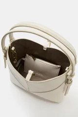 Bolso tipo mini shopper de diseño estructurado en color beige, con forma de V en la parte superior y asa de mano fija. Presenta costuras visibles y detalles metálicos en los laterales.