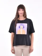Remera negra de corte clásico con estampado frontal cuadrado que ilustra a una persona con cabello violeta y chaqueta naranja.