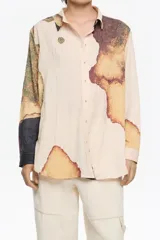 Camisa de corte recto con cuello camisero y botones frontales. La tela presenta un efecto arrugado o texturizado en tono beige claro, con grandes bloques de estampado abstracto en tonos tierra (marrón, ocre, verde oliva) que simulan manchas o texturas orgánicas. Las mangas son largas y una de ellas tiene un detalle en negro liso en el puño.