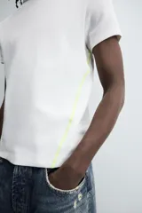 Camiseta de corte relajado, color gris claro, confeccionada en tejido con estructura waffle. Presenta cuello redondo y manga corta, con costuras decorativas en contraste color dorado que recorren los laterales y los hombros.