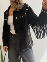 Chaqueta de efecto gamuza en color chocolate, con diseño de flecos en el pecho y mangas. Presenta detalles de tachas metálicas plateadas a lo largo de los hombros, solapas y bolsillos. Cuenta con cierre frontal de botones y forro interior.