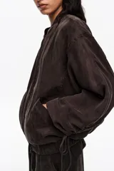 Chaqueta corta de corte bomber color marrón oscuro, confeccionada en mezcla de lyocell y viscosa. Presenta cierre frontal con cremallera, cuello alto, mangas largas con detalles fruncidos en los puños y bolsillos laterales.