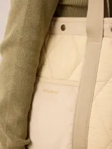 Bolso tipo tote de diseño acolchado con patrón de rombos, confeccionado en poliamida negra. Presenta un bolsillo frontal central de pana con el logo de la marca bordado en dorado, detalles de ribetes en pana negra y botones metálicos dorados en la parte superior. Incluye correas de hombro resistentes y forro interior de algodón a cuadros.