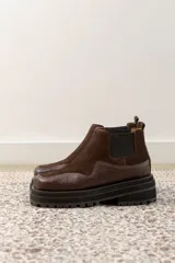 Botas tipo chelsea de caña baja, confeccionadas en una combinación de cuero gamuzado y cuero texturizado en tono marrón oscuro. Presentan una suela track de plataforma gruesa y elásticos laterales negros.